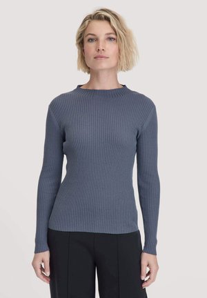 Blonde Frau mit kurzen gewellten Haaren, die einen enganliegenden grauen Langarmpullover aus Rippstrick und schwarze Hosen trägt, steht frontal vor einem schlichten Hintergrund.