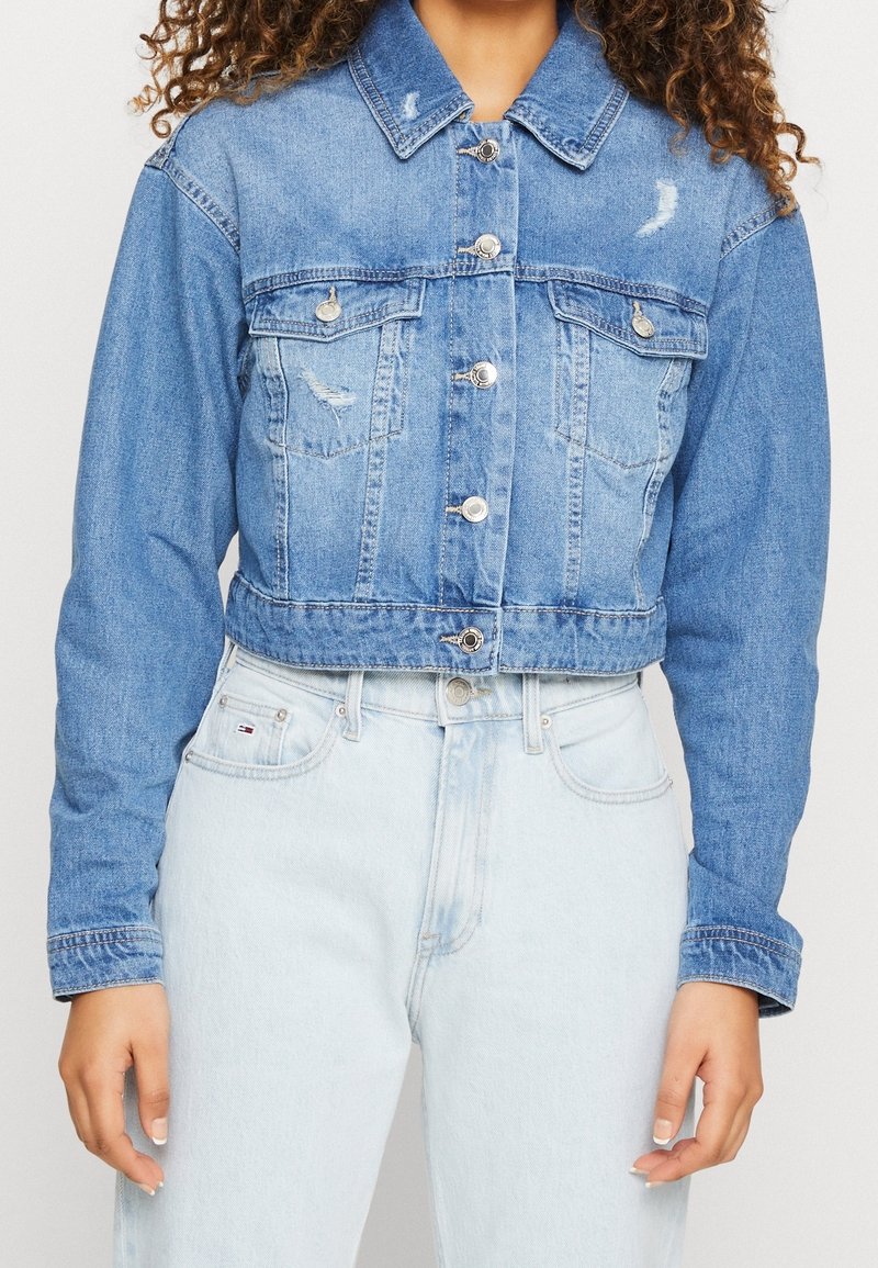 Mujer que lleva una chaqueta de mezclilla azul recortada con cierres de botón y detalles desgastados, combinada con unos pantalones vaqueros de cintura alta de color azul claro.