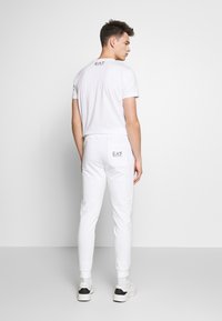 Λευκό βαμβακερό t-shirt και ταιριαστά joggers, με ελαστικές μανσέτες, λογότυπο στην πλάτη και σχέδιο σε στενή εφαρμογή. Φοριούνται με λευκά αθλητικά παπούτσια.
