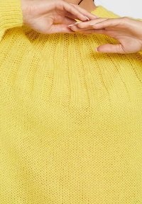 Yellow knitted sweater with a loose fit, featuring a ribbed neckline and soft, textured fabric. Hands adjusting the sweater are visible.