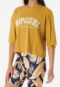 Musztardowa żółta krótka koszulka z napisem "RIP CURL" w białym kolorze; zestawiona z granatowymi szortami z jasnym kwiatowym wzorem. Miękka tkanina, luźny krój.