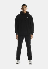 Juodas džemperis su siūlėmis, papuoštas mažu baltu logotipu. Sujungtas su juodomis kelnėmis ir baltomis sportinėmis avalynėmis, rodomas prieš baltą foną.