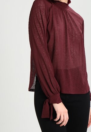 Chemisier bordeaux avec un col haut, en tissu texturé, avec des détails froncés et des poignets noués ; présente des fentes latérales pour un style supplémentaire.