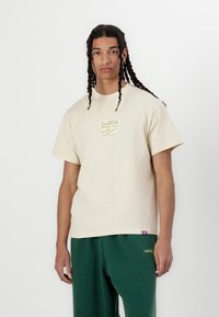 Lys beige bomull t-skjorte med et firkantet grafisk design i grønn og svart tekst. Avslappet passform, korte ermer og et lilla merkelapp ved kanten.
