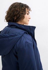 Giacca imbottita blu navy con collo alto e cappuccio attaccato, dotata di zip frontale e una texture liscia e lucida.