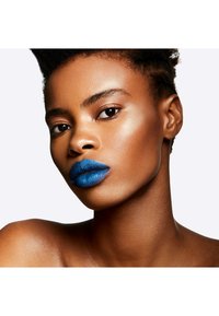 Gros plan d'un visage avec des lèvres texturées bleu profond, une peau lisse bronzée, des pommettes mises en valeur et des cheveux noirs coiffés près du cuir chevelu.