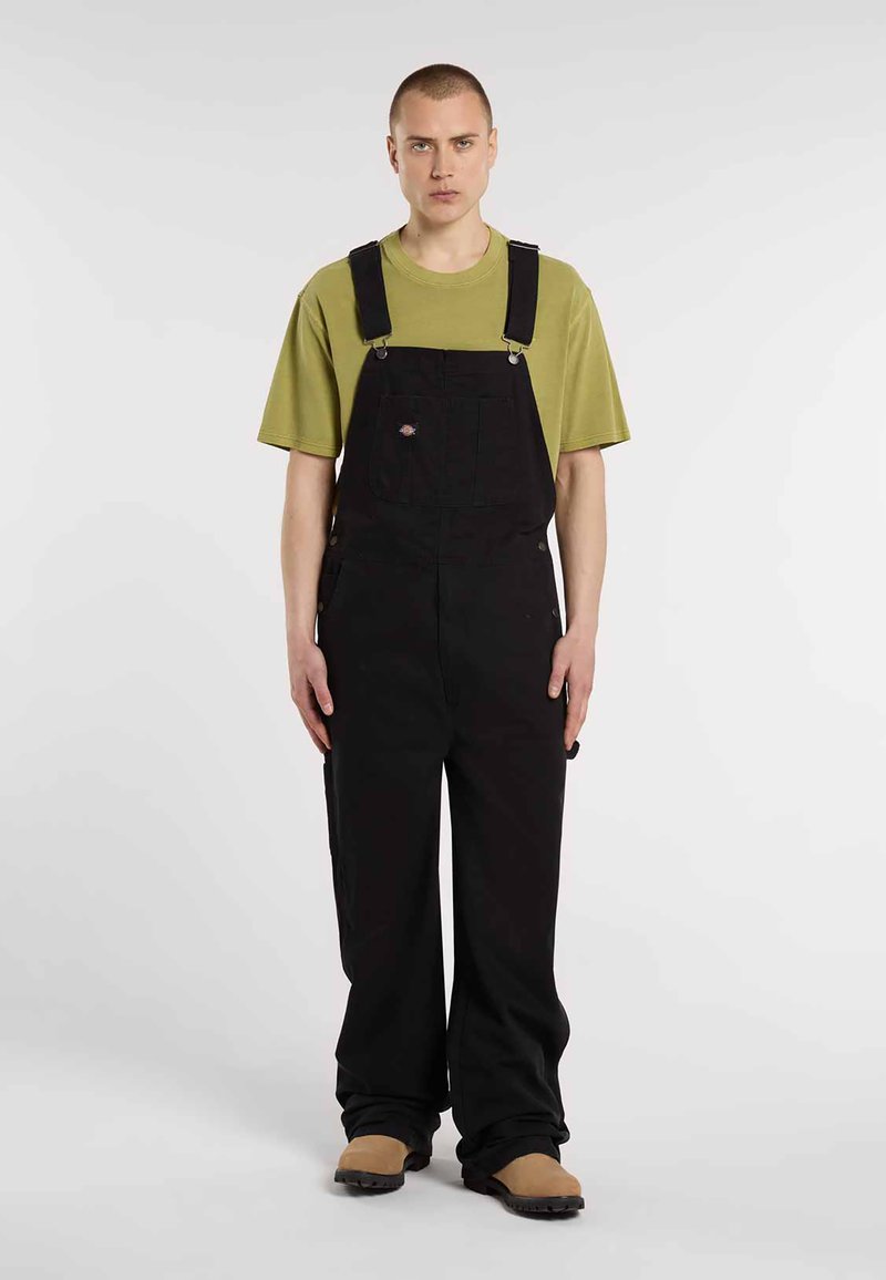 Sorte overalls med justerbare stropper, featuring en brystlomme og løse ben. Parret med en grøn t-shirt med korte ærmer og brune fodtøj.