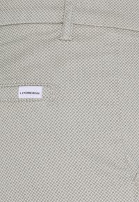 Gros plan sur un tissu beige clair texturé avec de petits motifs en losange et une étiquette blanche portant la mention "LINDBERGH" cousue près d'une poche.