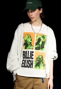 Hvid bomuld t-shirt med fed sort tekst "BILLIE EILISH" og grøn-orange grafiske billeder. Sammen med en grå zip-up hoodie og brune bukser.