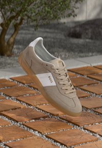 Beżowe zamszowe sneakersy z białymi skórzanymi akcentami, sznurowana konstrukcja, gumowa podeszwa oraz złoty detal z logo. Unoszą się nad teksturowaną powierzchnią płytek.