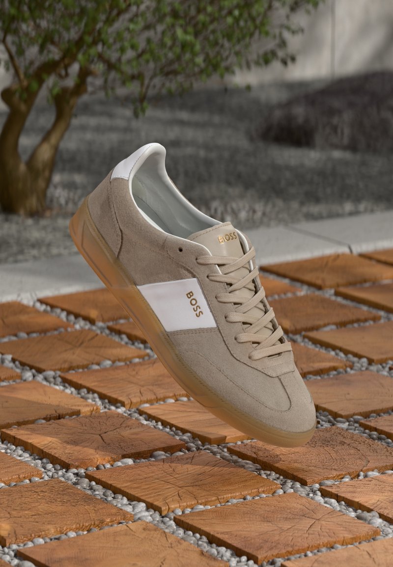 Beżowe zamszowe sneakersy z białymi skórzanymi akcentami, sznurowana konstrukcja, gumowa podeszwa oraz złoty detal z logo. Unoszą się nad teksturowaną powierzchnią płytek.