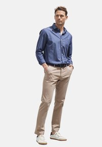 Blaues Hemd mit Knöpfen aus gewebtem Stoff, kombiniert mit beigen Chinos und weißen Sneakers mit braunen Akzenten. Das Model steht mit den Händen in den Taschen.