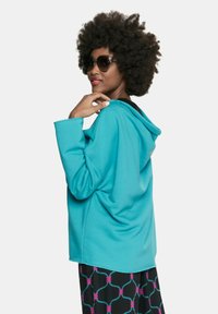 Turkusowa bluza z kapturem o miękkiej fakturze, z czarnym wykończeniem i luźnym krojem. W zestawie z wzorzystymi czarnymi i różowymi spodniami.