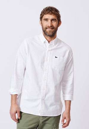 Mann mit braunen lockigen Haaren und Bart, trägt ein weißes langärmeliges Hemd und grüne Hosen, lächelt leicht vor einem schlichten Hintergrund.