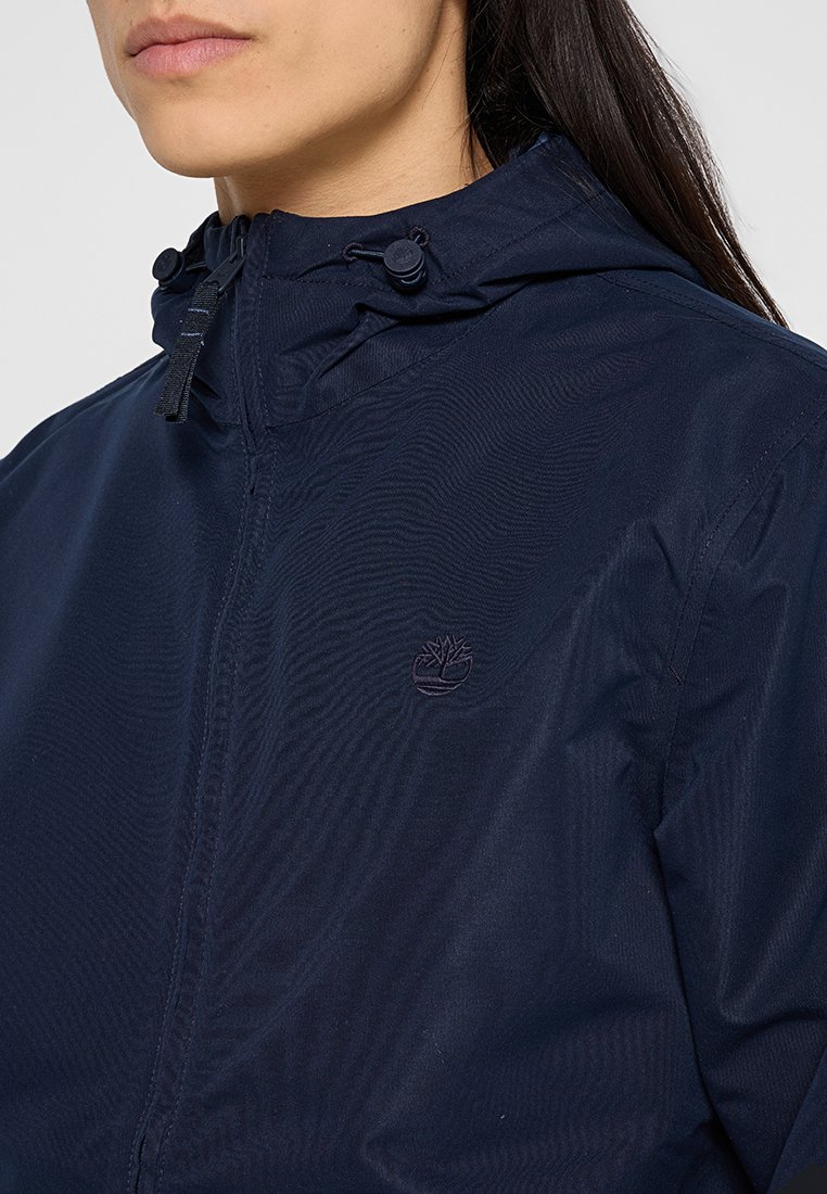 Granatowa kurtka z kapturem, wykonana z lekkiego materiału, z suwakiem i sznurkiem do regulacji, z dyskretnym haftowanym logo na klatce piersiowej.