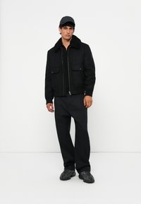 Chaqueta bomber negra con cuello de piel sintética, dos bolsillos frontales y cremallera. Combinada con pantalones oscuros y zapatillas negras texturizadas.