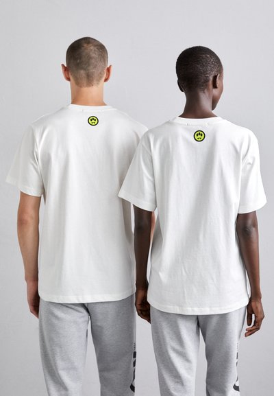 Białe bawełniane t-shirty z krótkim rękawem, z okrągłym zielonym logo uśmiechu na plecach. Noszone z szarymi dresami.