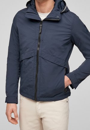 Mężczyzna w granatowej kurtce z kapturem zapinanej na zamek z przodu oraz w jasnobeżowych spodniach, z jedną ręką w kieszeni, na tle jednolitego szarego tła.