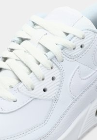 Zbliżenie na biały sneaker z przeszyciami, grubymi białymi sznurówkami i teksturowaną podeszwą z subtelnym logo na bocznej części.