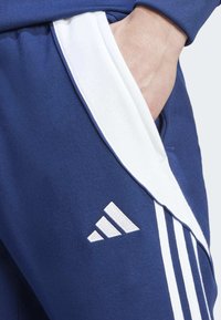 Jūrinės mėlynos sportinės kelnės su baltomis šoninėmis juostelėmis ir trimis baltomis juostelėmis. Yra kišenė, glotni tekstūra ir Adidas logotipo detalė ant šlaunies.