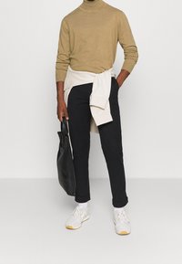 Maglione beige a collo alto, maglione crema legato in vita, pantaloni neri a sigaretta, borsa tote nera, sneakers bianche con suola beige chiaro.