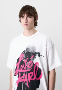 Fehér, oversized póló, amelynek feltűnő grafikája egy alakot ábrázol, mellette a "Love Karl" felirattal élénk rózsaszín színben. Rövid ujjú és puha tapintású.