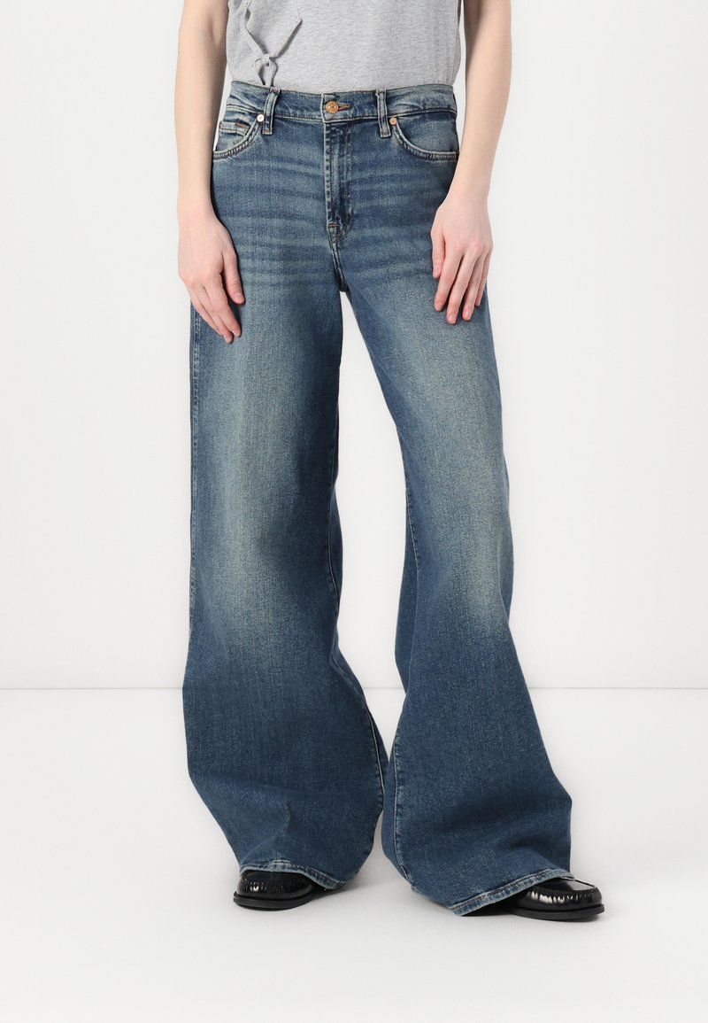 Světle modré oversized zvonové džíny s vybledlým vzhledem, vysokým pasem a designem s pěti kapsami. V kombinaci s šedým tričkem a černými botami.