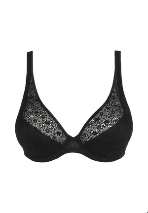 Femme portant un ensemble assorti de soutien-gorge et culotte en dentelle noire, debout et face à l’avant sur un fond gris uni.