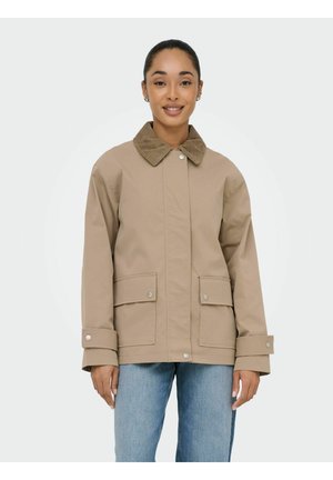 Femeie zâmbitoare purtând o geacă bej cu guler din catifea, buzunare cu capse și curele la manșete, asortată cu blugi albaștri deschiși.