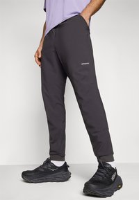 Donkergrijze, taps toelopende sportbroeken met een gladde textuur, voorzien van een logo op de dij en geribbelde enkelmanchetten. Gecombineerd met zwarte sneakers.