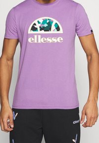 Lila t-shirt med kort ärm som har en färgglad camouflagegrafik och "ellesse"-logotyp i krämfärg. Tillverkad av bomull med en standardpassform.