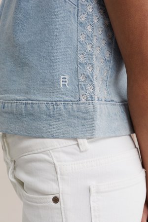 Hellblaue Jeansjacke mit weißem, besticktem Blumenmuster an der Seite, kombiniert mit weißer Hose am Oberkörper einer Person.