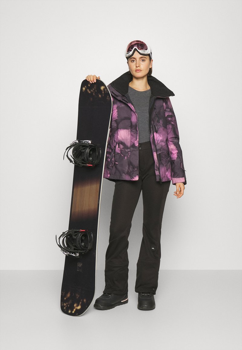 Snowboard i narciarz w fioletowej kurtce w kwiatowy wzór, szarej koszulce, czarnych spodniach i okularach przeciwsłonecznych, trzymający ciemny snowboard z wiązaniami.