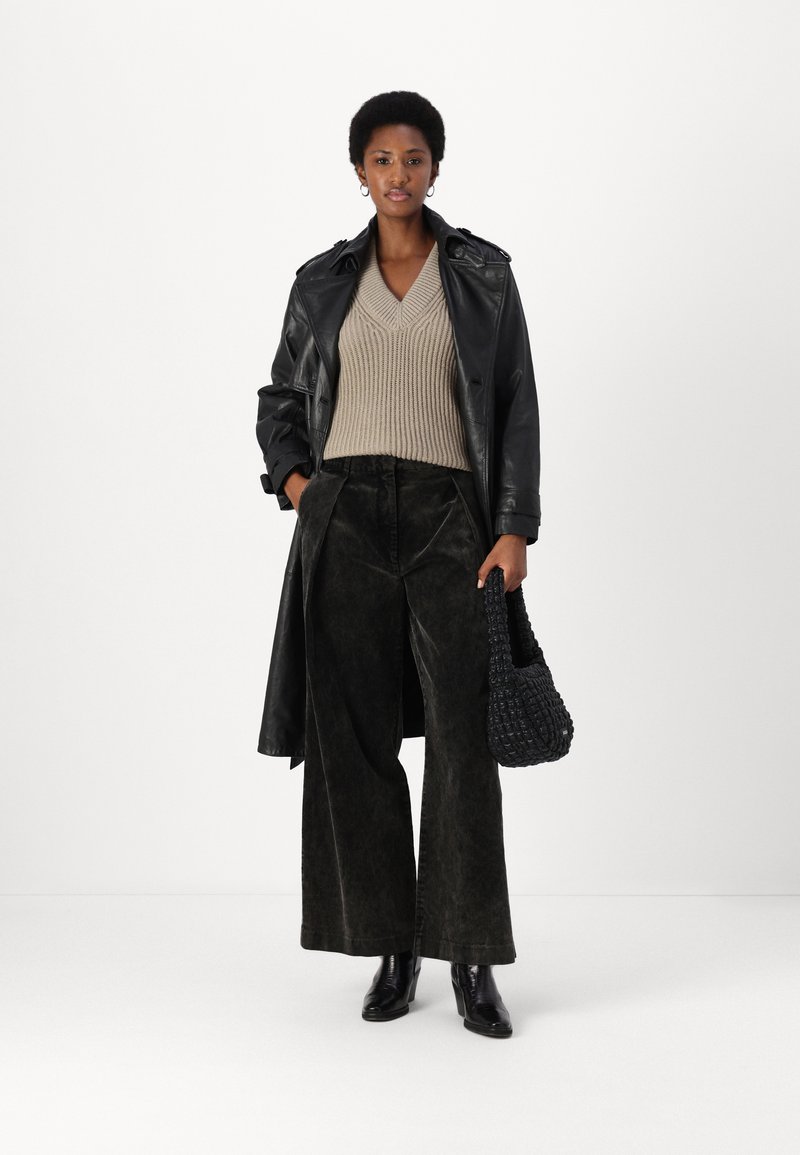 Schwarzer Leder-Trenchcoat, beiger gerippter Pullover, dunkel strukturierte weit geschnittene Hose, schwarze Absatzstiefel und eine gewebte schwarze Handtasche.