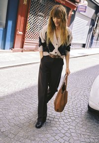 Femme aux longs cheveux portant un cardigan à motifs et un pantalon noir marche dans une rue pavée en tenant un sac fourre-tout marron.