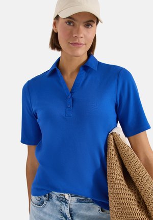 Jeune femme en polo bleu à manches courtes, casquette beige, tenant un sac marron texturé, debout devant un fond clair uni.