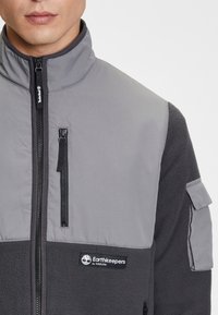 Grijze fleecevest met een hoge kraag, ritssluiting aan de voorkant, borstzak en contrasterende stofpanelen met een Earthkeepers-logo.