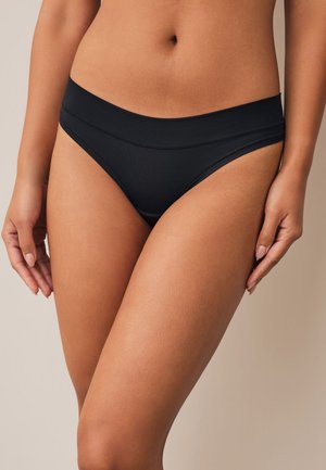 Kvinde iført sort bikinitrusser med mid-rise, stående mod en neutral baggrund med hænderne afslappet ved siden.