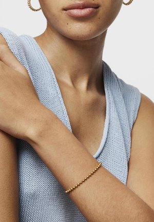 Pulsera de cuentas de oro llevada en la muñeca, combinada con una blusa sin mangas de punto azul claro. La piel tiene una textura suave, y la tela presenta un patrón texturizado.