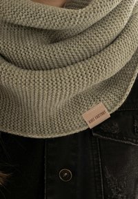 Sciarpa lavorata a maglia beige con una texture intrecciata avvolta attorno al collo sopra una giacca di denim nera con un bottone e l'etichetta "Knit Factory" ben visibile.