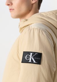 Bežna kapuca z mat teksturo, s črnim patchom in belim logotipom na rokavu. Preprost in funkcionalen dizajn.