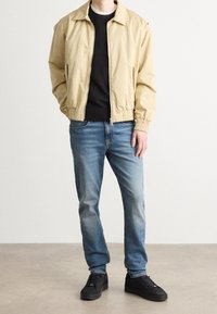 Light beige bomber jacket with elastic cuffs, worn over a black sweater, paired with light blue jeans and black sneakers.