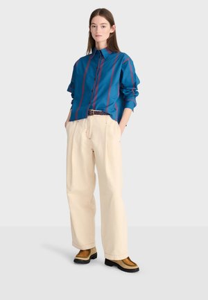 Jeune femme debout, les mains dans les poches, portant une chemise rayée bleue, un pantalon large crème et des chaussures marron sur un fond clair.