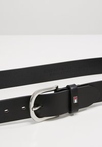 Svart läderbälte med en silverfärgad metallspänne, med en liten logotyp i röd och blå, samt sju justeringshål. Slät textur.