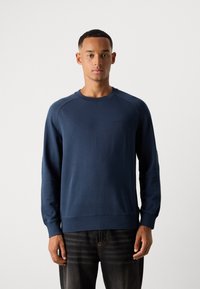 Ung mann iført en marineblå hettegenser og svarte jeans, stående rett mot en lys, ensfarget bakgrunn.