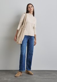 Beiger Rollkragensweater, helle Denim-Hose mit weitem Bein und beigefarbene Schuhe. Hält eine flauschige, beigefarbene Tote-Tasche. Glattes Material, lässiges Design.