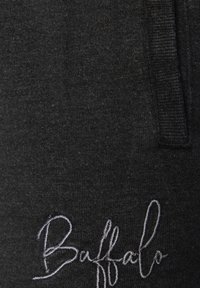 Gros plan sur un tissu noir avec une texture douce, présentant un logo "Buffalo" brodé en blanc. Le design inclut une taille visible et des détails de couture.