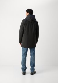 Parka negra con capucha, de textura suave, con un respaldo ligeramente más largo. Llevada sobre jeans azules, mostrada contra un fondo neutro.