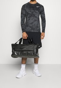 Hombre con camisa de manga larga negra con estampado y pantalones cortos sosteniendo un bolso de deporte negro de Nike, vistiendo calcetines blancos y zapatillas deportivas blancas frente a un fondo liso.