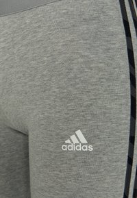 Aproximação do tecido cinza de vestuário desportivo Adidas com riscas laterais pretas e o logótipo branco da Adidas na área da coxa.